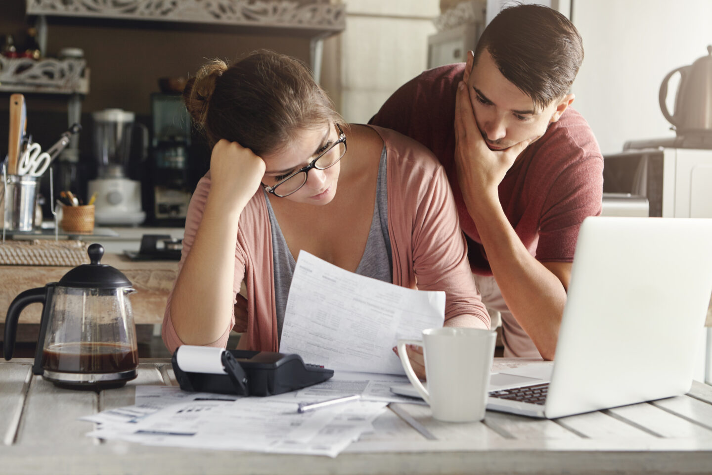 Young Stressed Caucasian Couple Facing Financials Troubles, Sitting At Kitchen Table With Papers, Calculator And Laptop Computer And Reading Document From Bank, Looking Frustrated And Unhappy