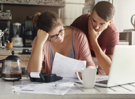 Young Stressed Caucasian Couple Facing Financials Troubles, Sitting At Kitchen Table With Papers, Calculator And Laptop Computer And Reading Document From Bank, Looking Frustrated And Unhappy